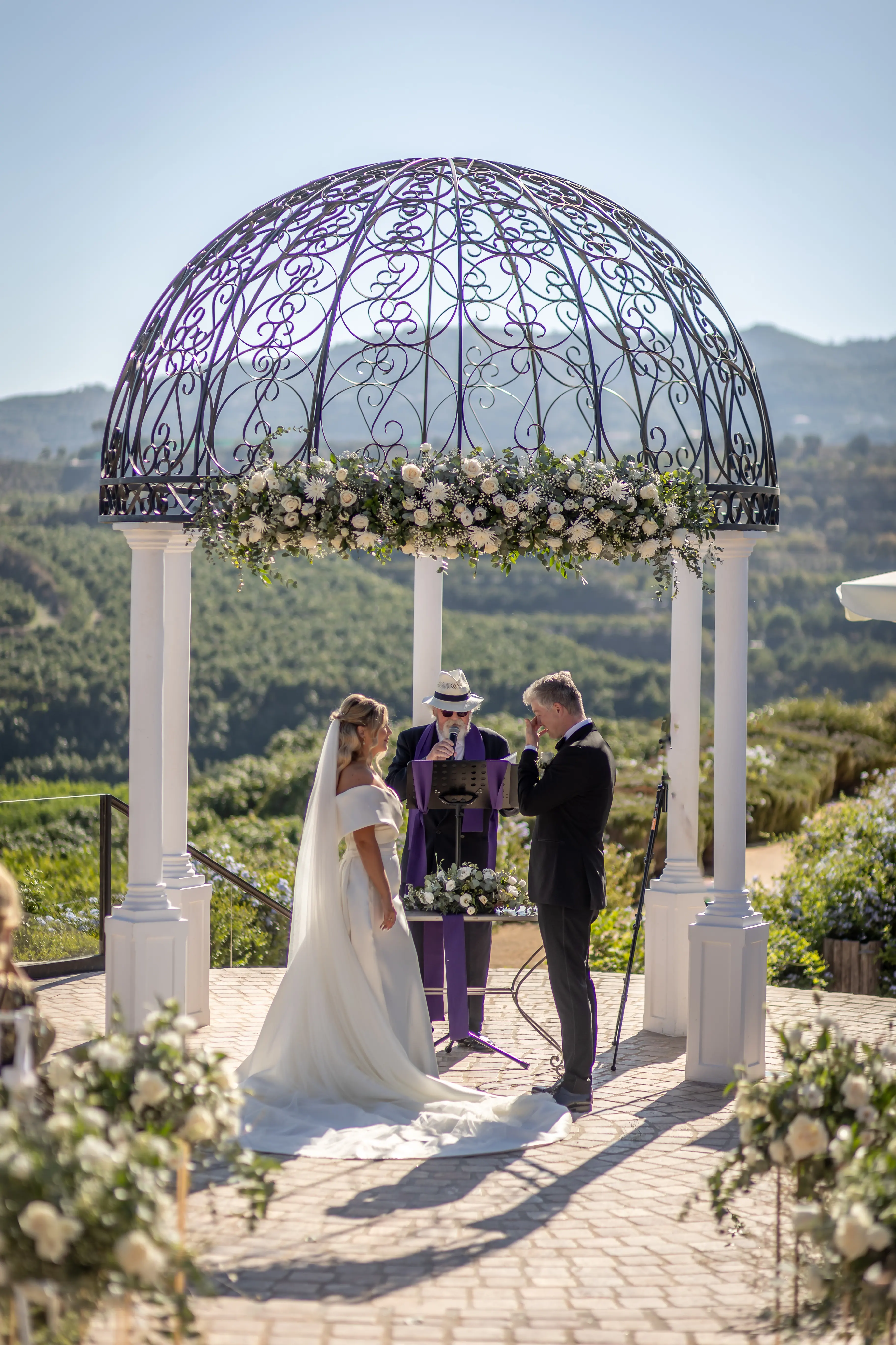 A wedding ceremony beneath a domed floral pergola overlooking the hills