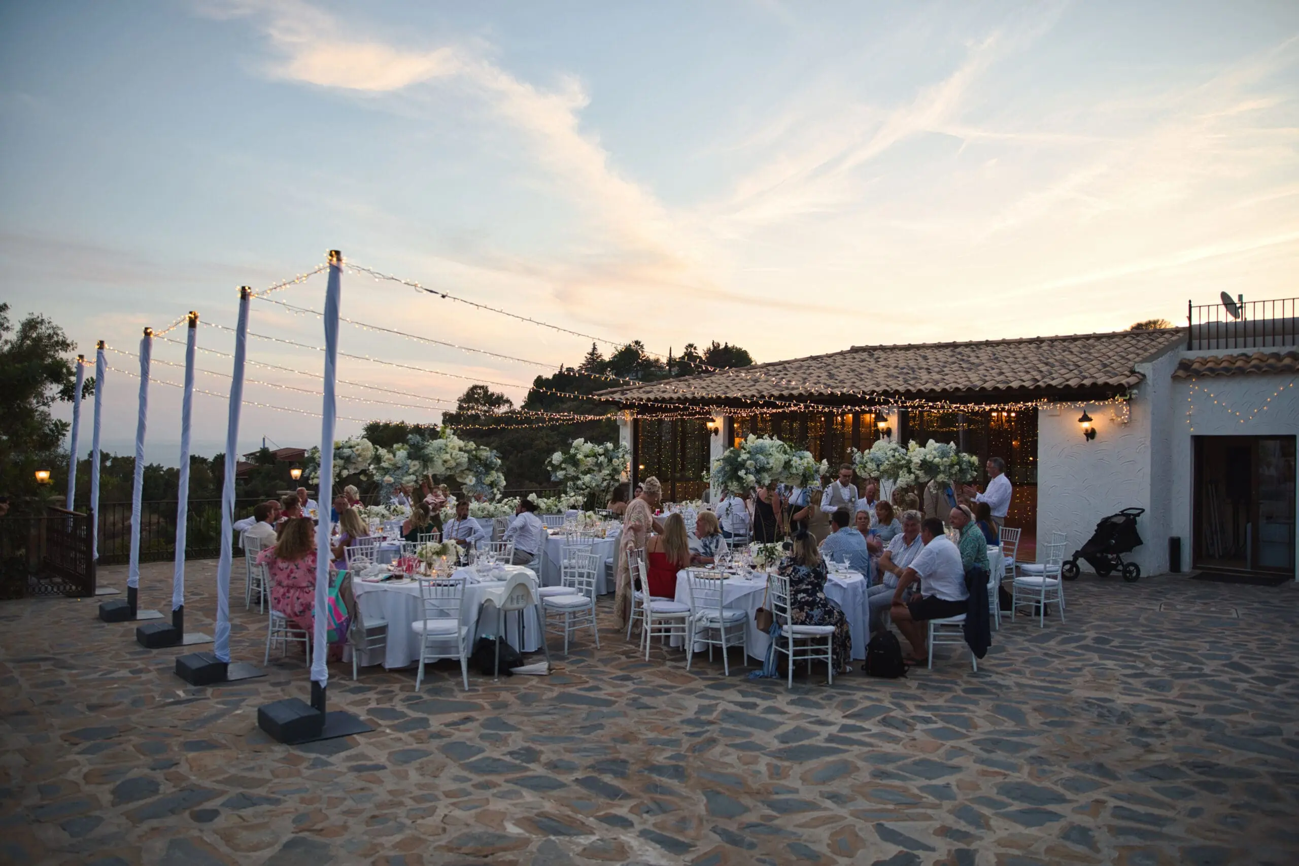 Guests seated at long tables outside a white villa as the sun sets over the hills